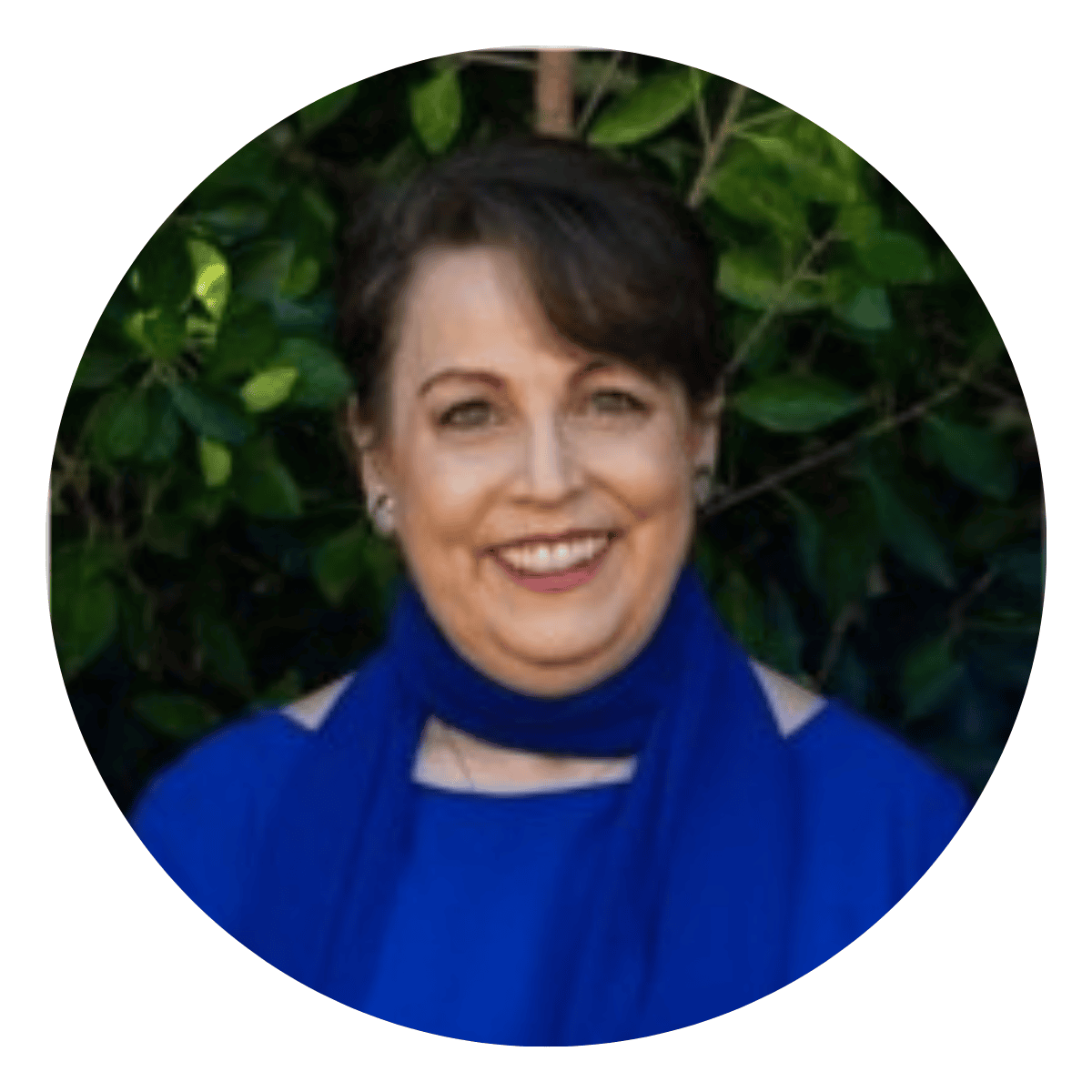 Headshot of a white female mid-50s executive coaching client in a blue top and scarf in front of a dark tree