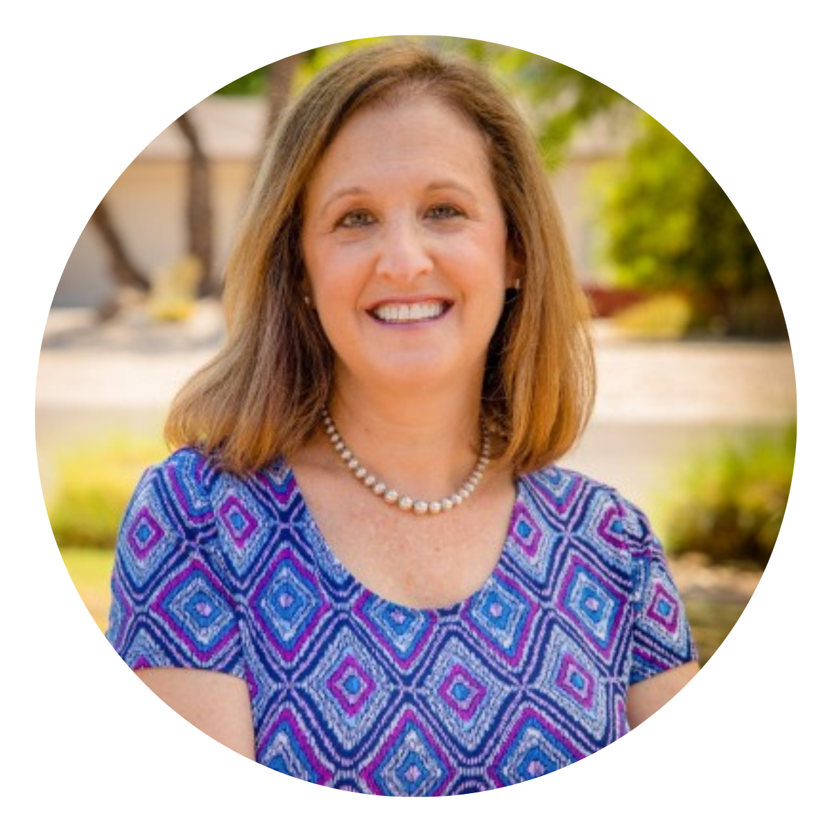 Headshot of a white female mid-50s executive coaching client in a patterned blue dress outside in the sunshine
